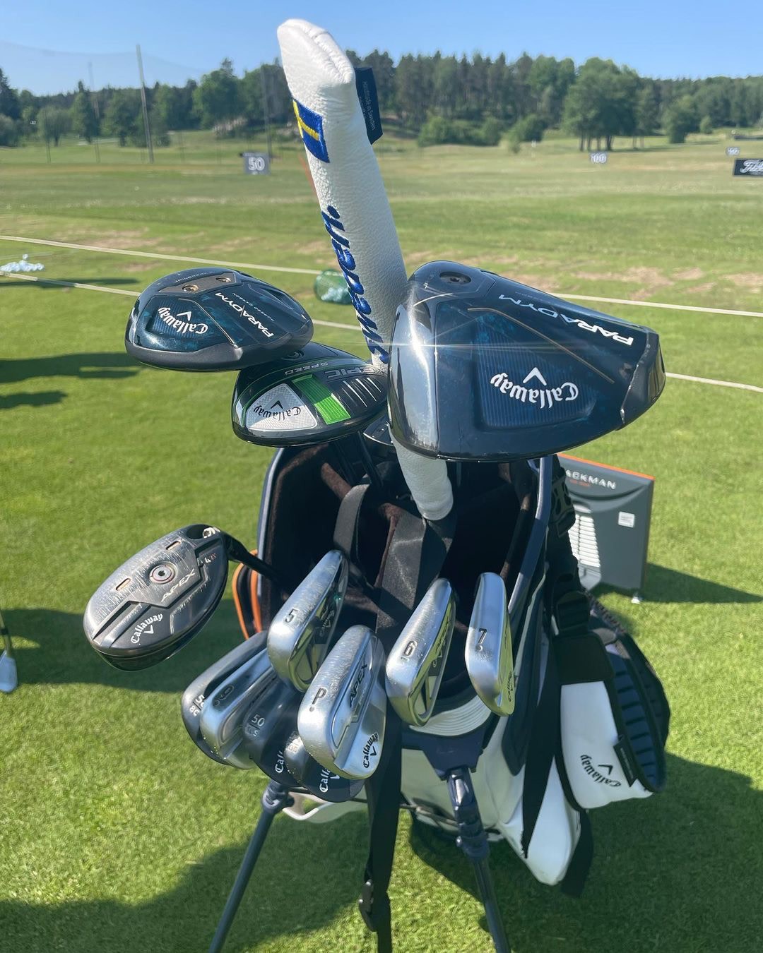 Golfbag med Callaway-klubbor står på en driving range med grönt gräs och träd i bakgrunden under en klarblå himmel.