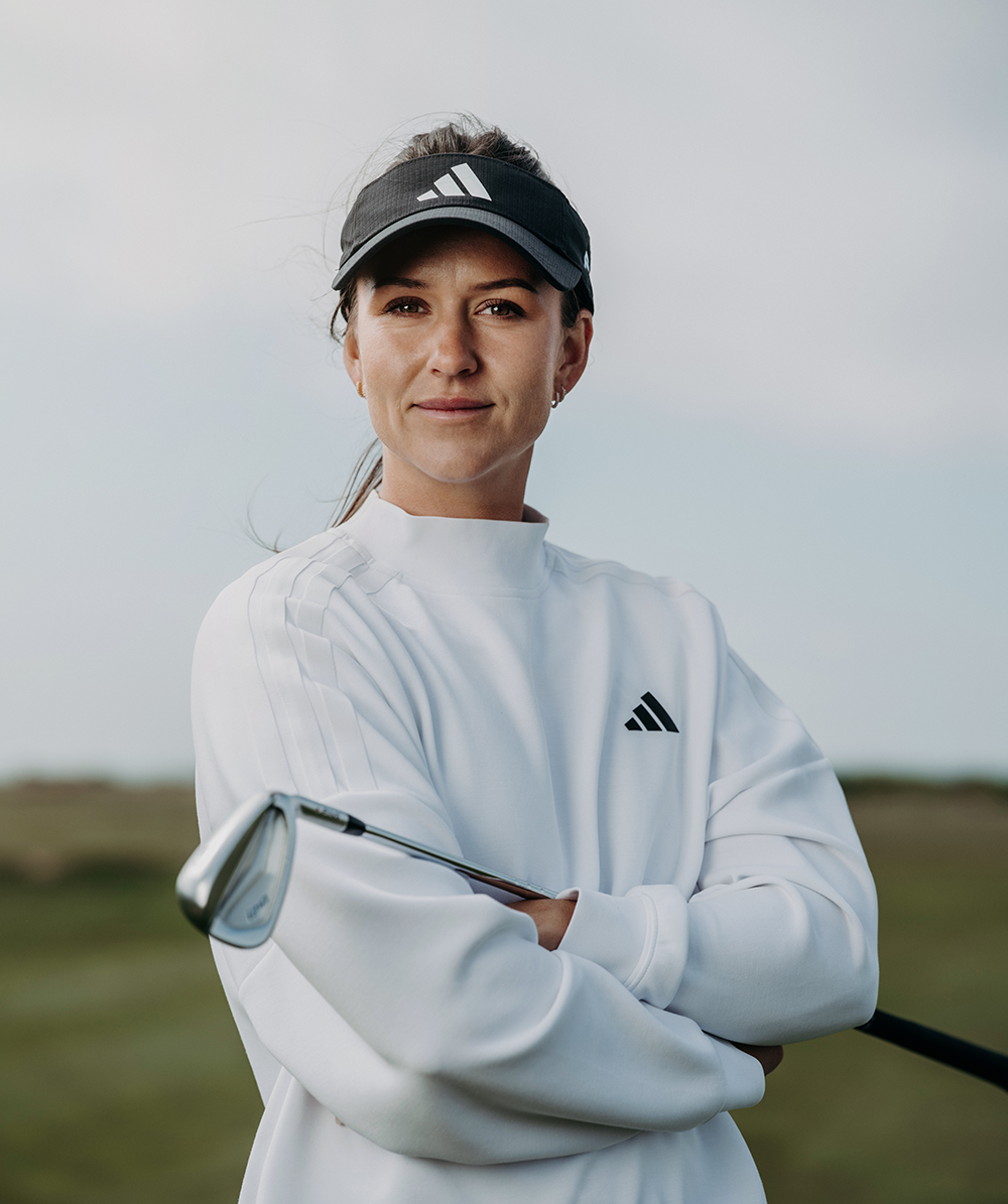 Kvinna i vit Adidas-tröja och svart visir håller en golfklubba med armarna i kors på en golfbana.