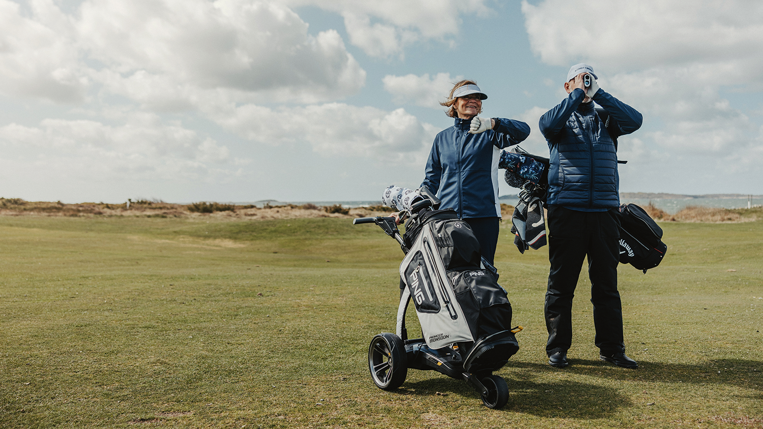Två golfare står på en golfbana; en kvinna ler bredvid en golfvagn och en man tittar genom en avståndsmätare.
