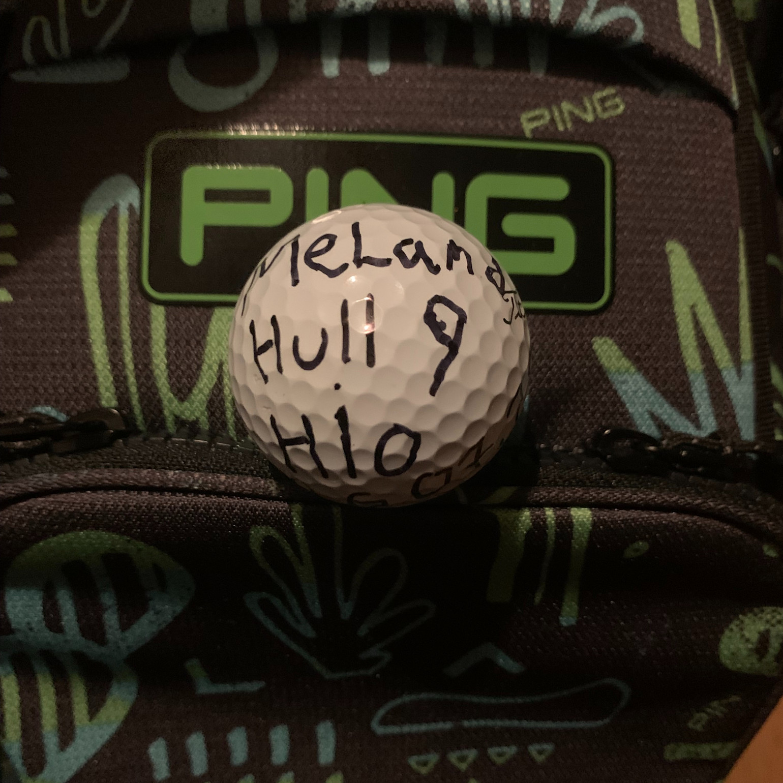 En golfbold med håndskrevet tekst "Melander, Hul 9, HIO" ligger på en golfbag med PING-logo i baggrunden.