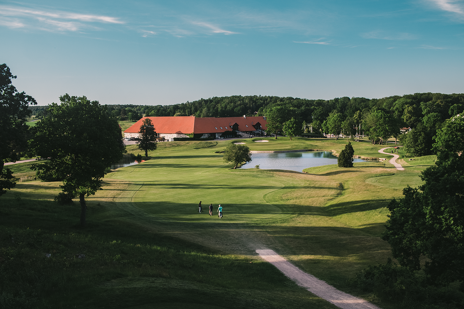 Tre personer går på en grön golfbana med träd, damm och en stor byggnad med rött tak i bakgrunden under klar himmel.
