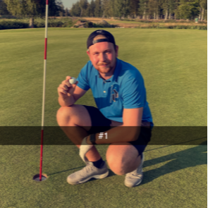 Mand i blå polo og kasket sidder på hug ved et golfhul, holder en golfbold og smiler på en golfbane.