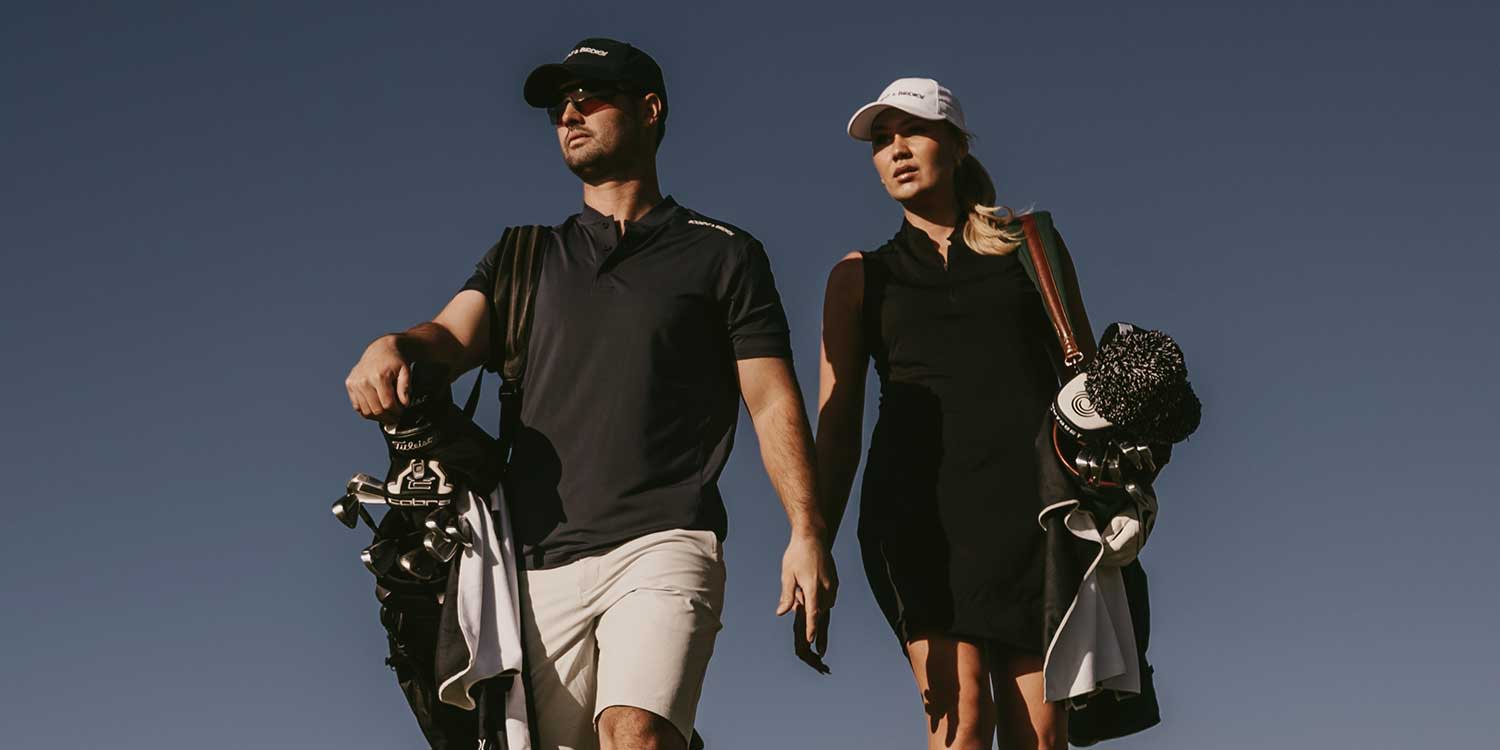 Two golfers walk side by side with golf bags over their shoulders under a clear blue sky.