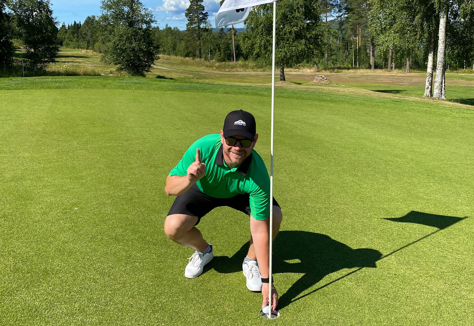 Mand i grøn poloshirt og sort kasket huker sig ved et golfhul, holder en golfbold og viser ét med fingeren på en solrig golfbane.