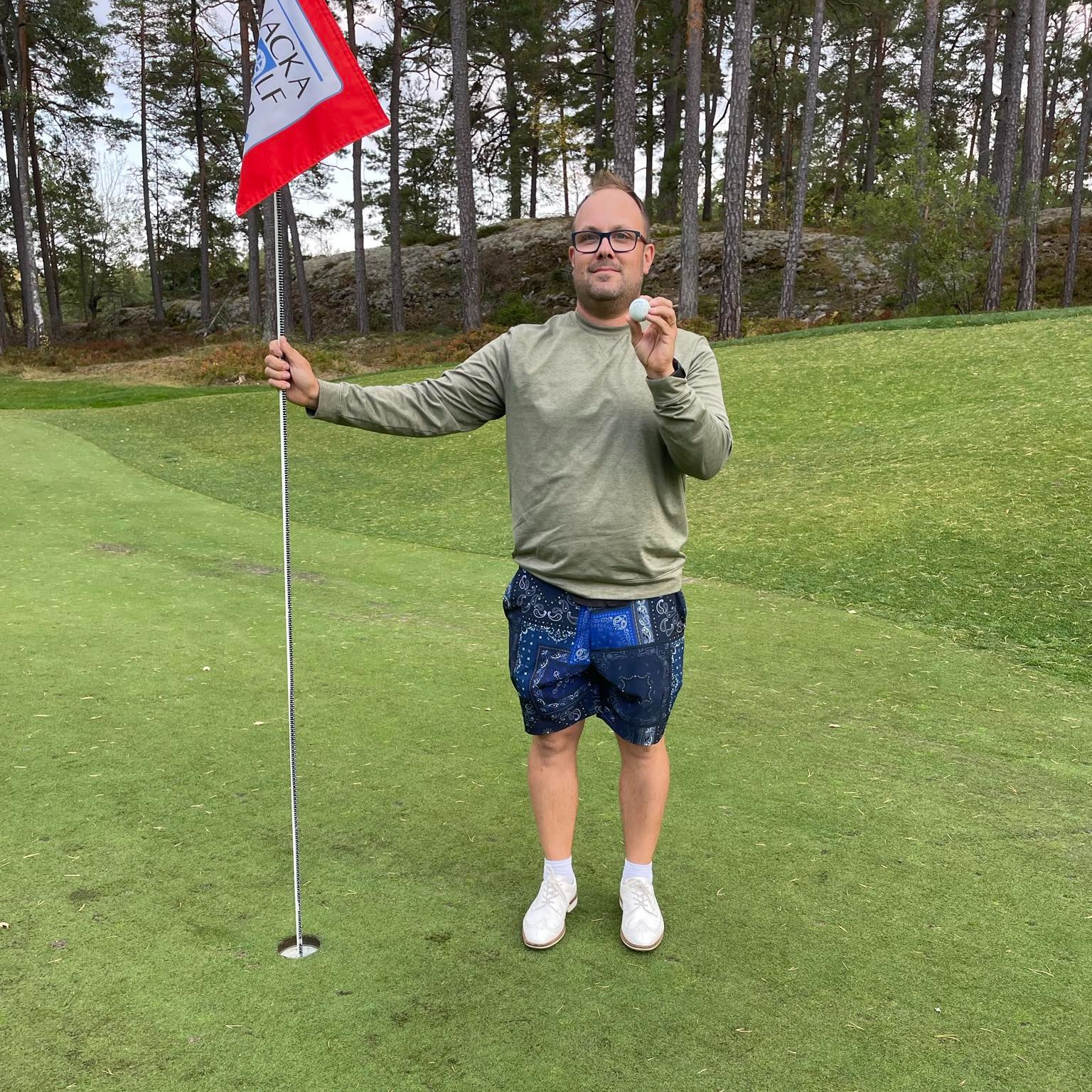 Man håller en golfboll och flaggstång på en golfbana, omgiven av gräs och träd i bakgrunden.