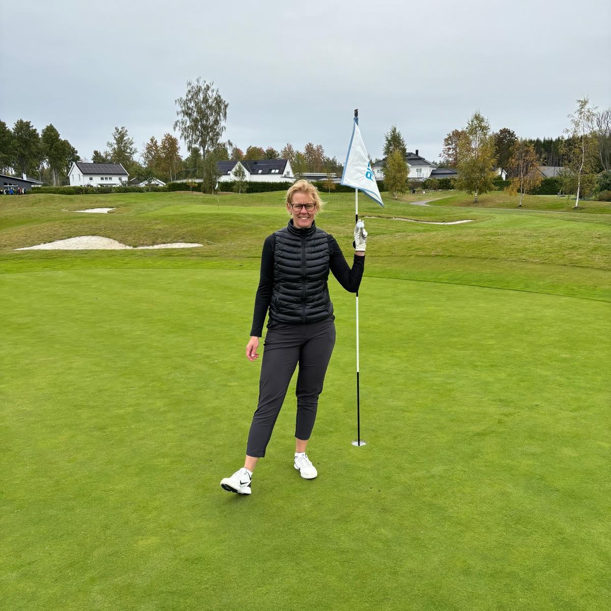 Kvinna står på en golfbana och håller i en flaggstång på greenen, med hus och träd i bakgrunden.