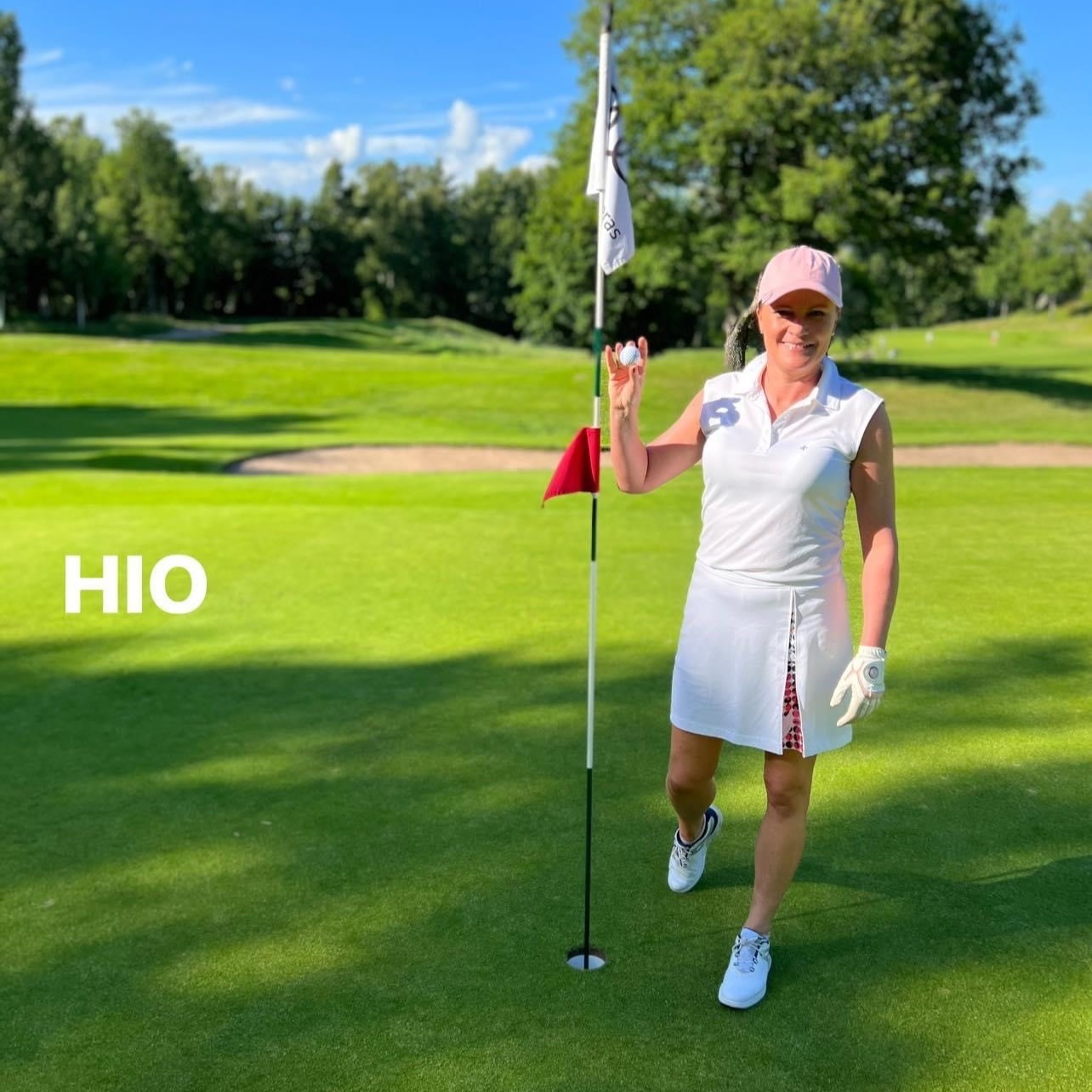 Kvinde i hvidt golfoutfit holder en golfbold op ved siden af hullet på greenen, med teksten "HIO" til venstre. Solrig dag på golfbanen.