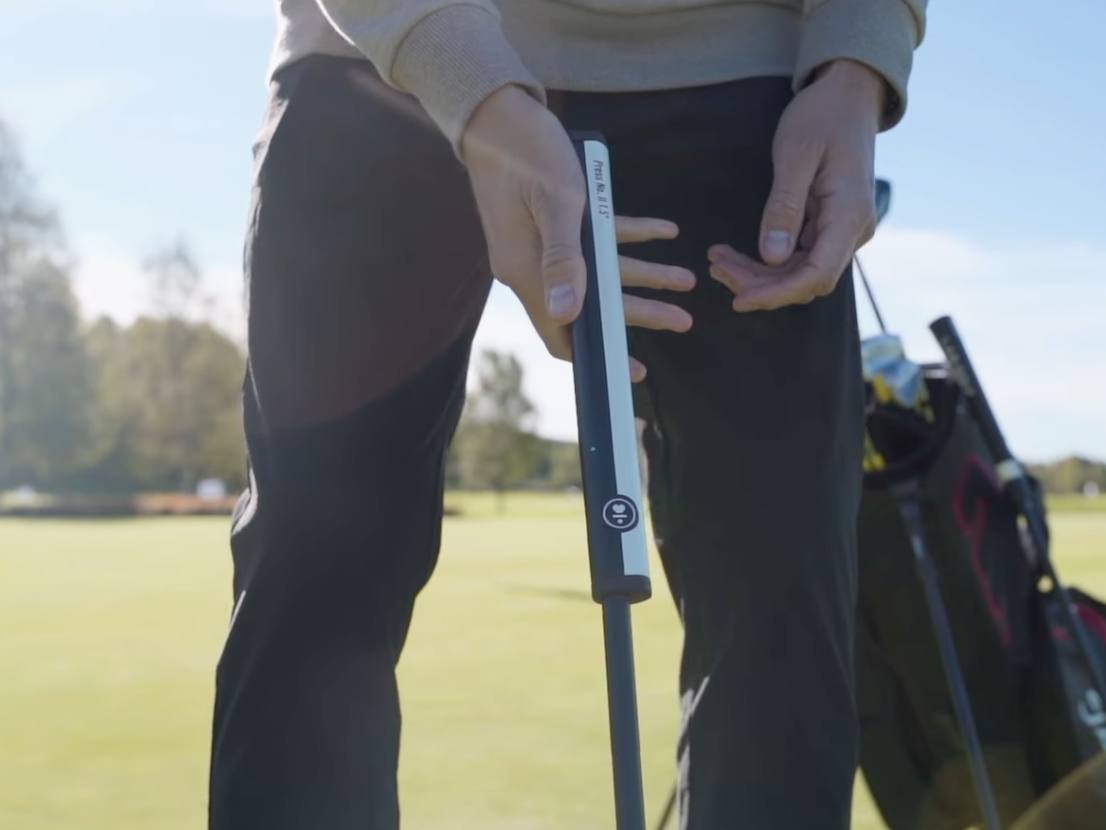 Eine Person hält einen Golfschlägerschaft auf einem Golfplatz, mit einer Golftasche im Hintergrund unter einem sonnigen Himmel.
