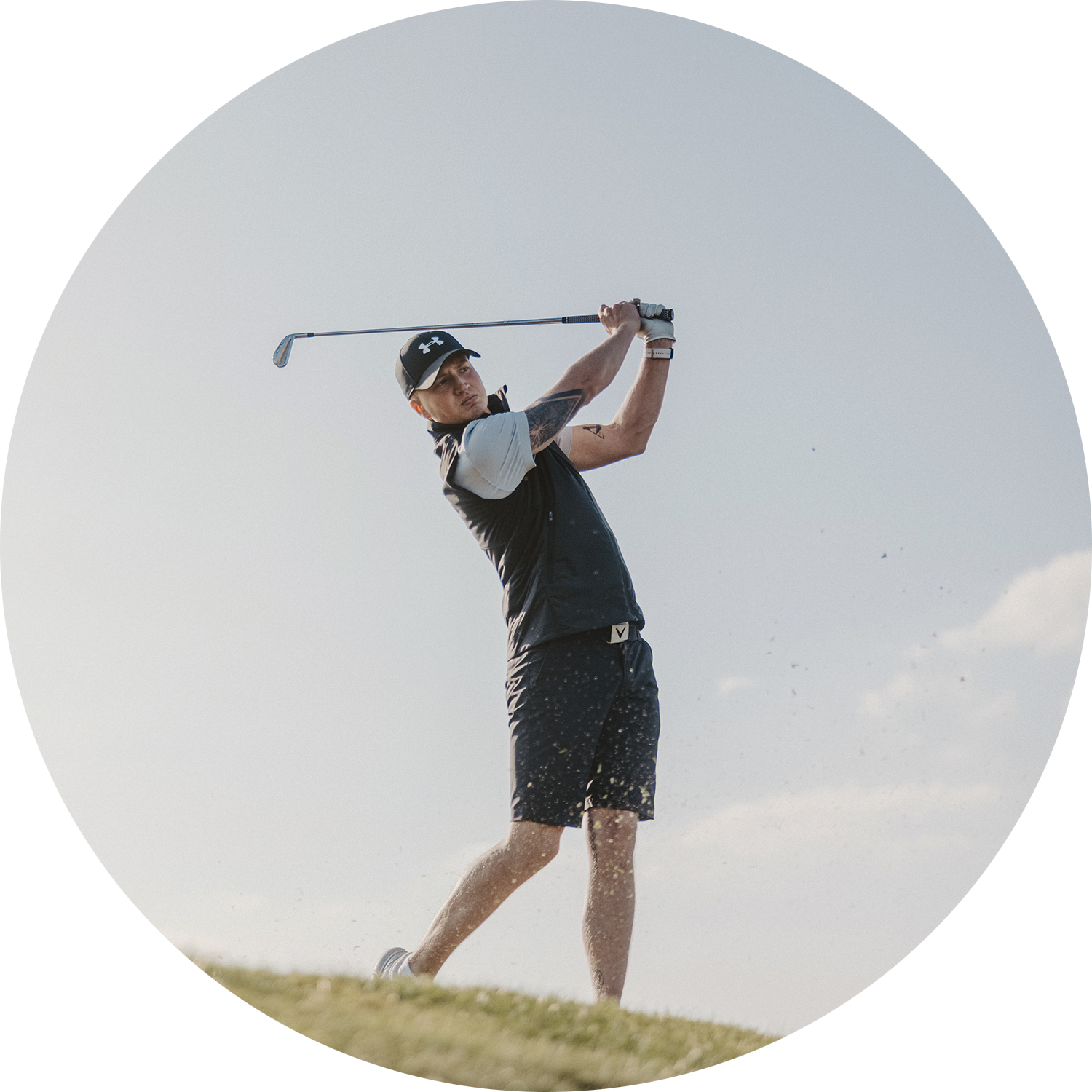 Mies lyö golflyöntiä golfkentällä, pukeutuneena urheiluvaatteisiin ja lippikseen kirkkaan taivaan alla.