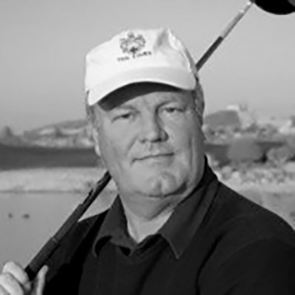 Man med golfklubba över axeln står utomhus, bär keps och tröja, med suddig bakgrund av landskap.