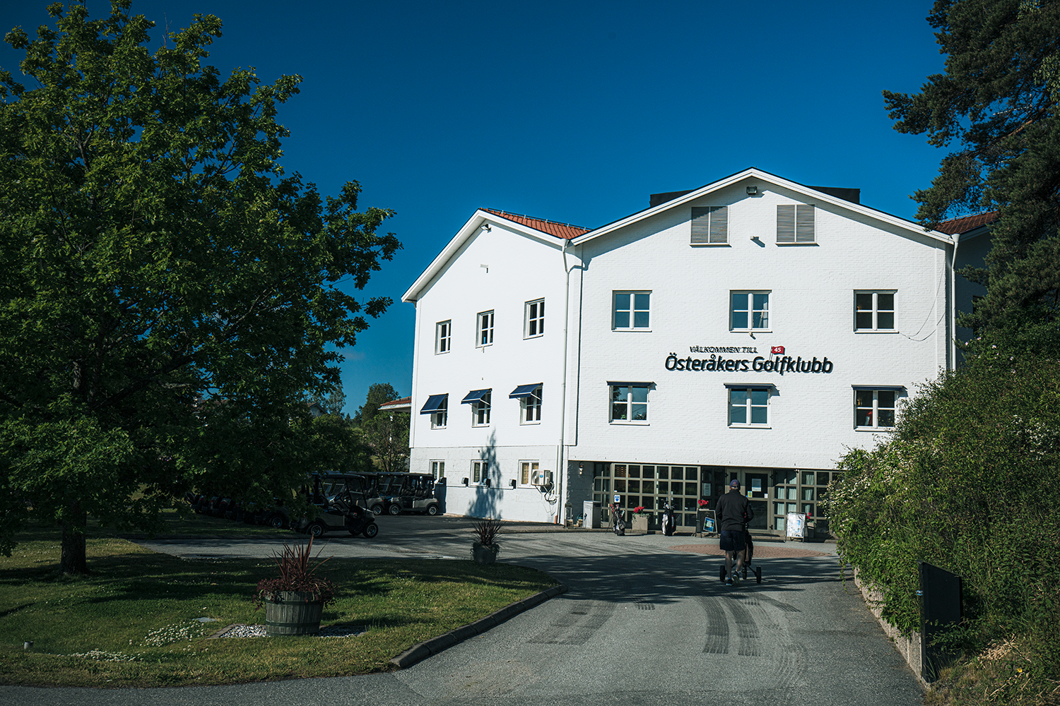 Vit byggnad med texten "Välkommen till Österåkers Golfklubb" på fasaden, omgiven av träd och golfbilar, klar blå himmel.