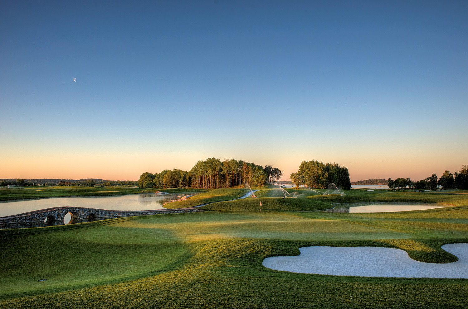 Golfbana med grönt gräs, sandbunkrar och vattenhinder, bevattningsspridare är igång i solnedgångsljus.