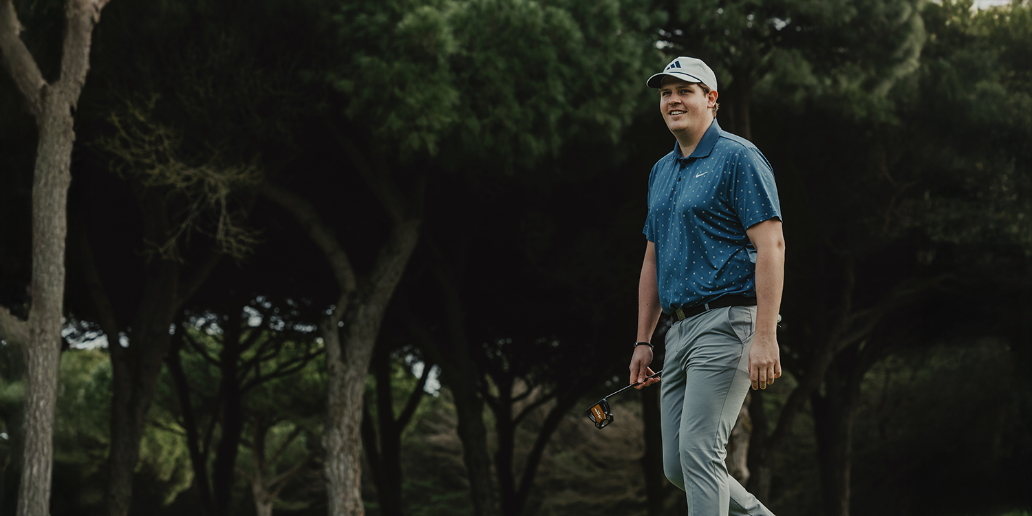 Man geht auf einem Golfplatz mit einem Golfschläger in der Hand, gekleidet in einem blauen Poloshirt, grauen Hosen und einer Kappe, umgeben von Bäumen.