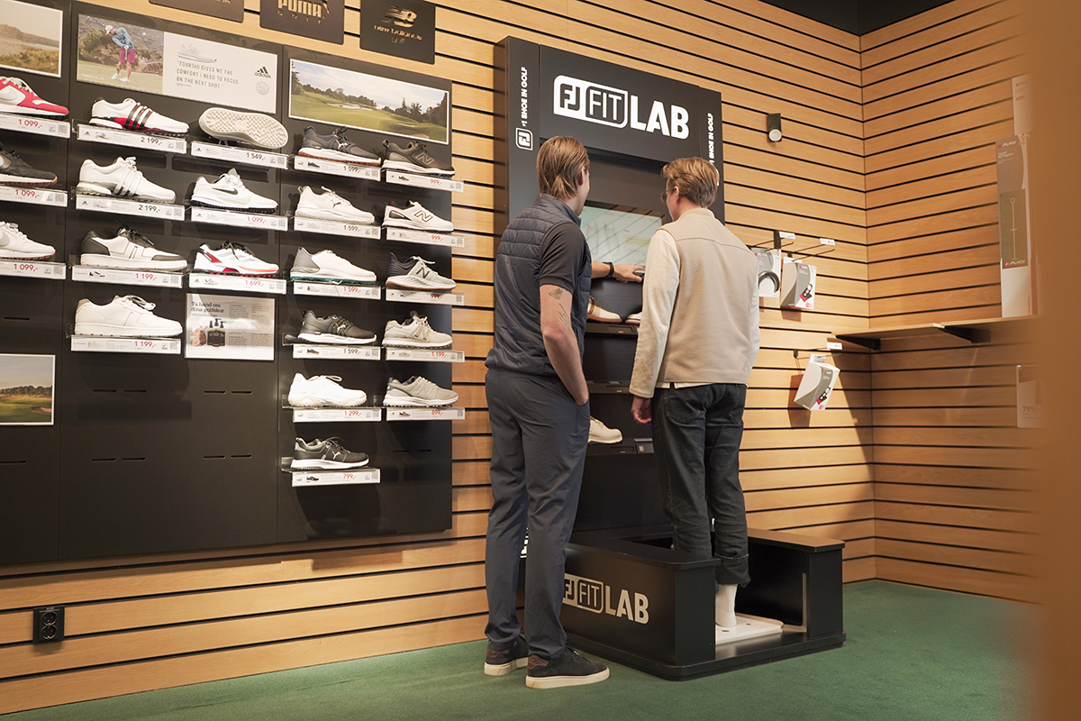 Två män står vid en FJ Fit Lab-station i en butik, en får hjälp att mäta sina fötter, omgivna av golvskor på väggen.