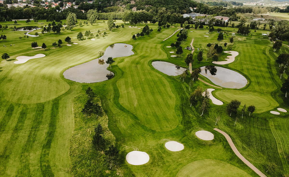 Flera golfbanor med gröna fairways, sandbunkrar och dammar omgivna av träd och gångvägar i ett öppet landskap.