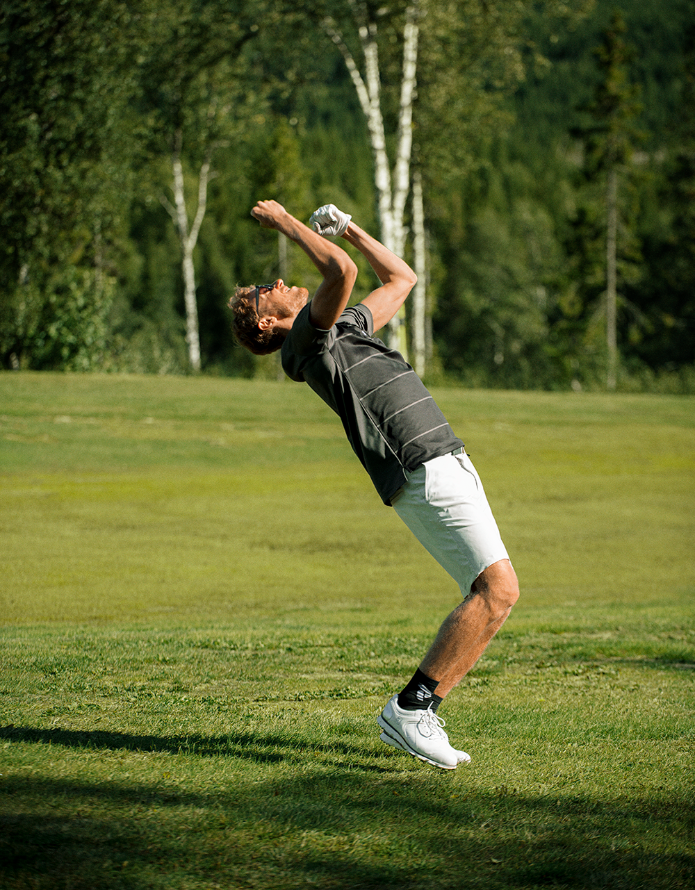 Man i golfkläder lutar sig bakåt och firar med armarna i luften på en golfbana omgiven av träd.