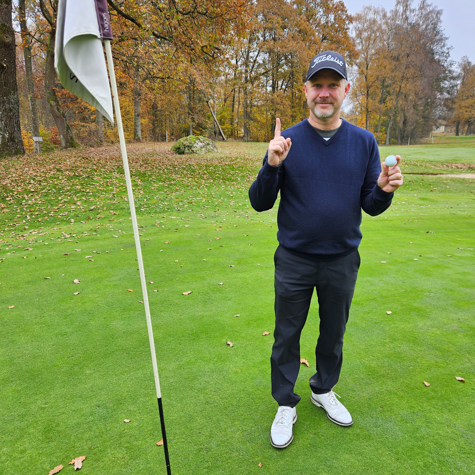 Man i golfkläder håller upp ett finger och en golfboll bredvid ett hål på en golfbana med höstfärgade träd i bakgrunden.