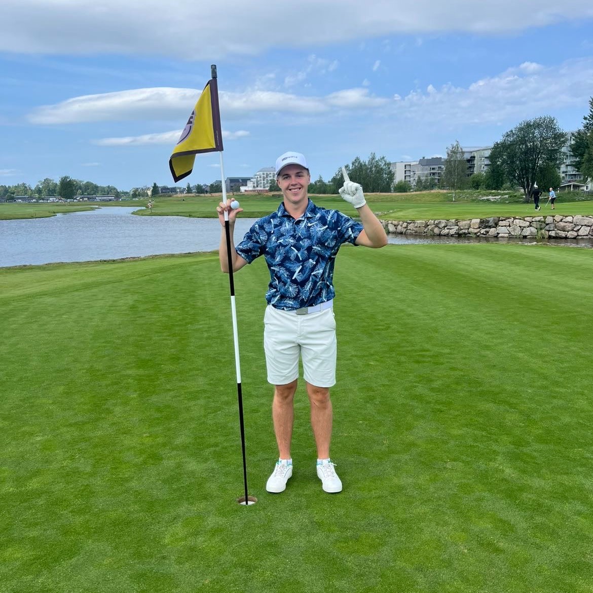 A man in golf attire is holding a flagstick and a golf ball on the green, smiling and making a victory gesture at a golf course with water and buildings in the background.