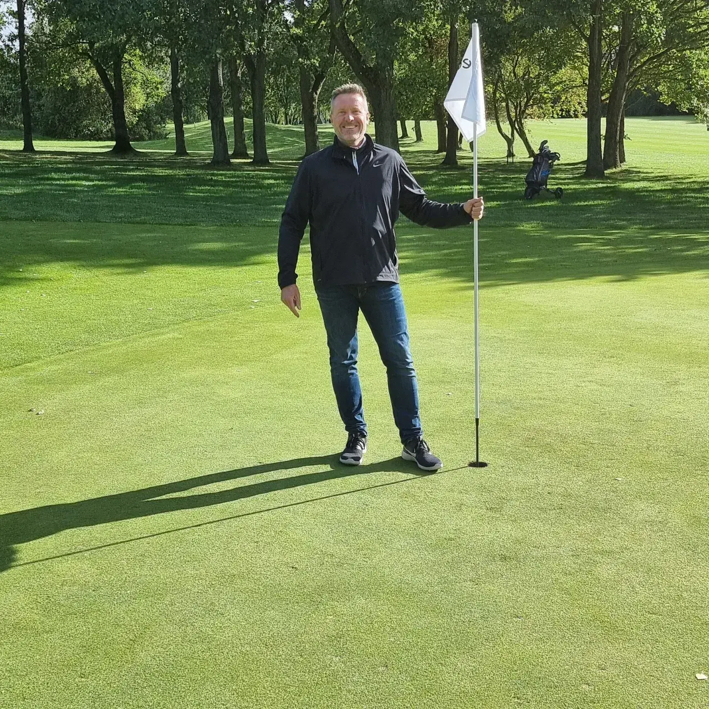 Man holder en golfflag ved hullet på en grøn golfbane med træer i baggrunden, og en golfvogn er synlig længere væk.