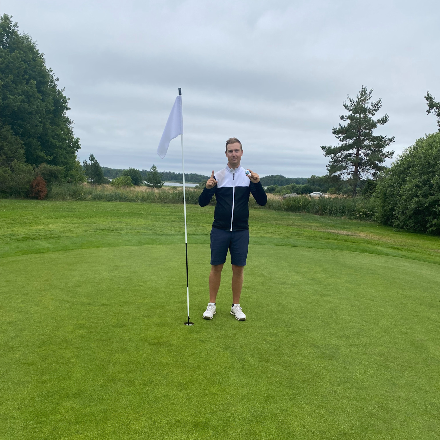 Man står ved siden af et golf-flag på en green, peger opad med begge hænder, omgivet af grønne planter og træer under en overskyet himmel.