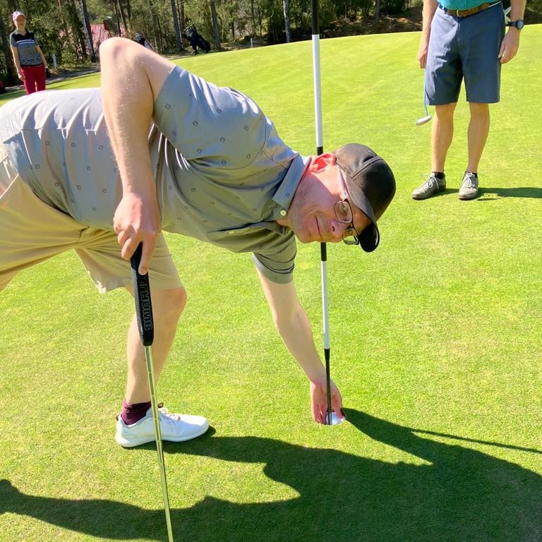 Man bøjer sig ned for at samle en golfbold op fra hullet på en golfbane, med to andre personer i baggrunden.