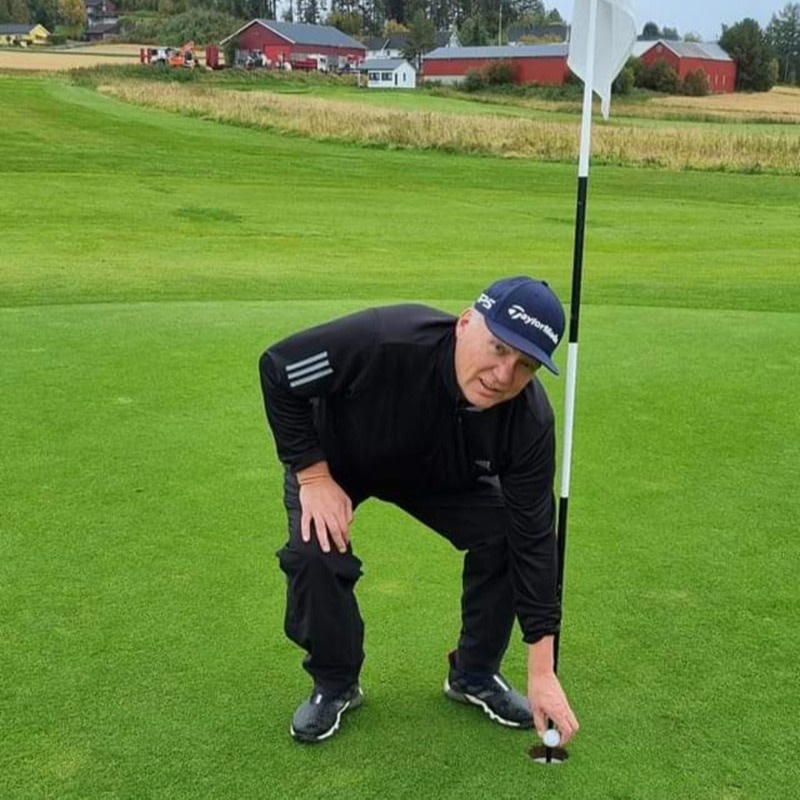 Mand iført sort golfbeklædning bøjer sig ned for at tage en golfbold op af hullet på en golfbane med landlig baggrund.