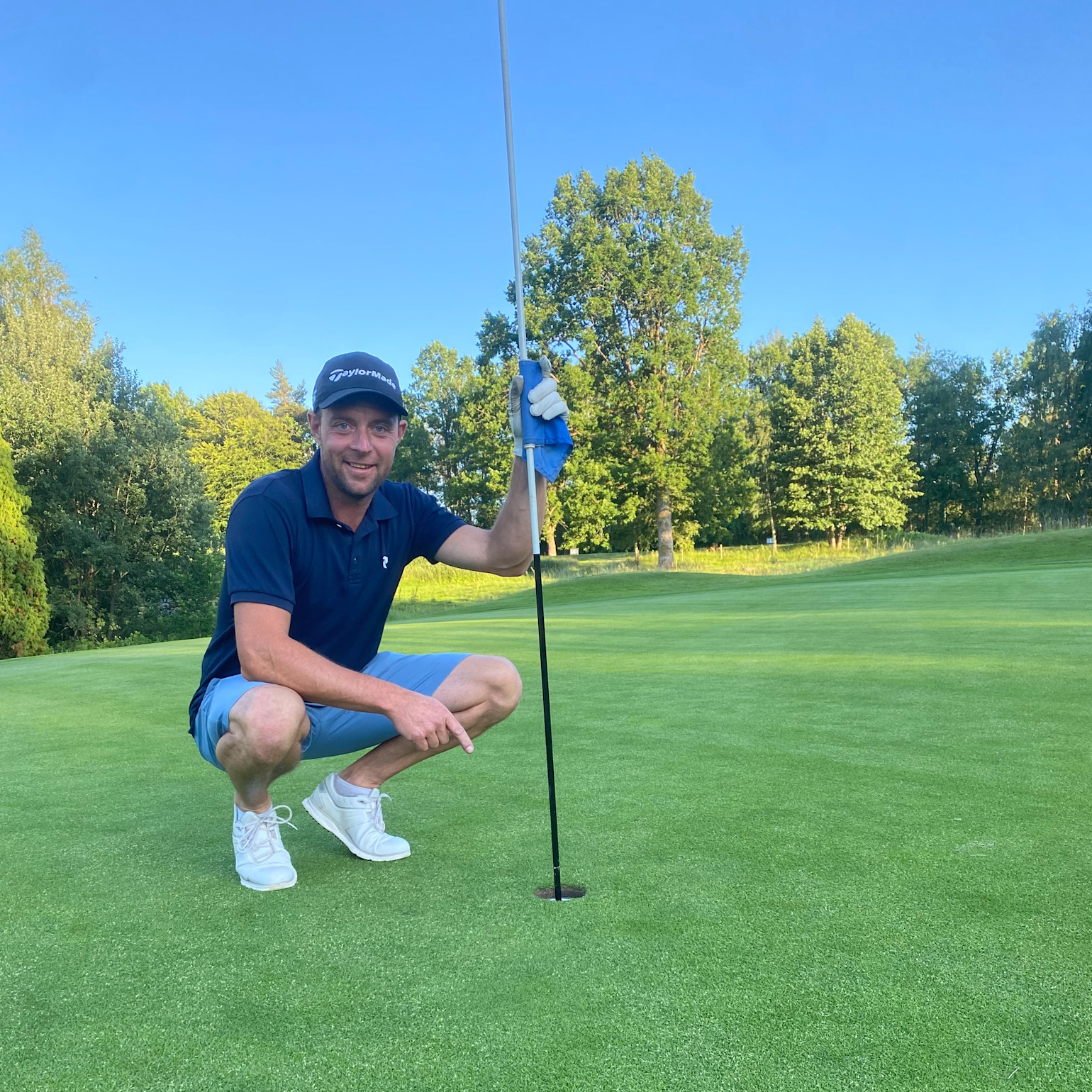 En mand iført golfbeklædning sidder på hug ved et golfhul og holder i flagstangen på en grøn golfbane med træer i baggrunden.