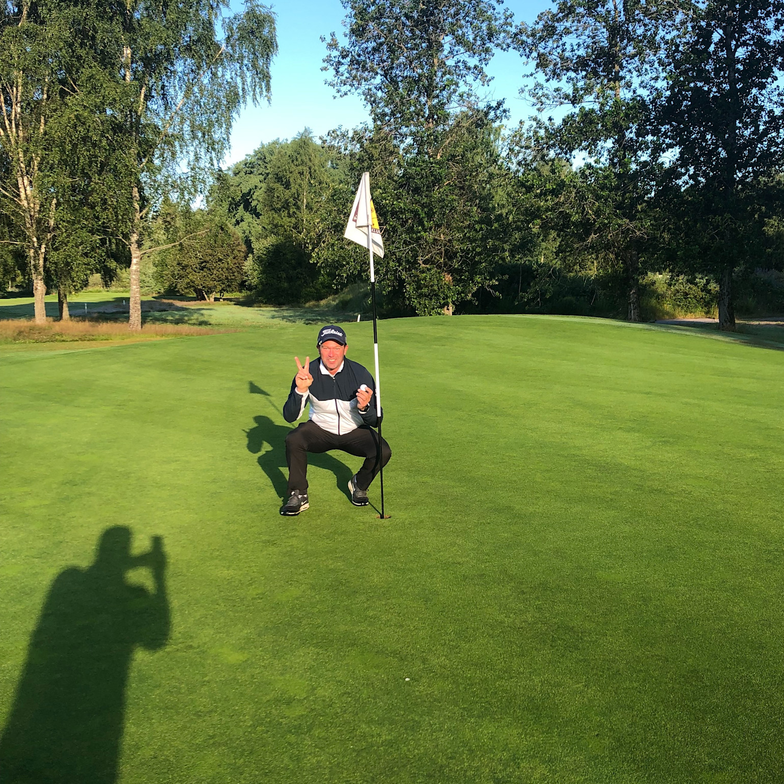 En person sidder på hug ved et golfhul med flag på en grøn golfbane, laver sejrtegn og er omgivet af træer i solskin.