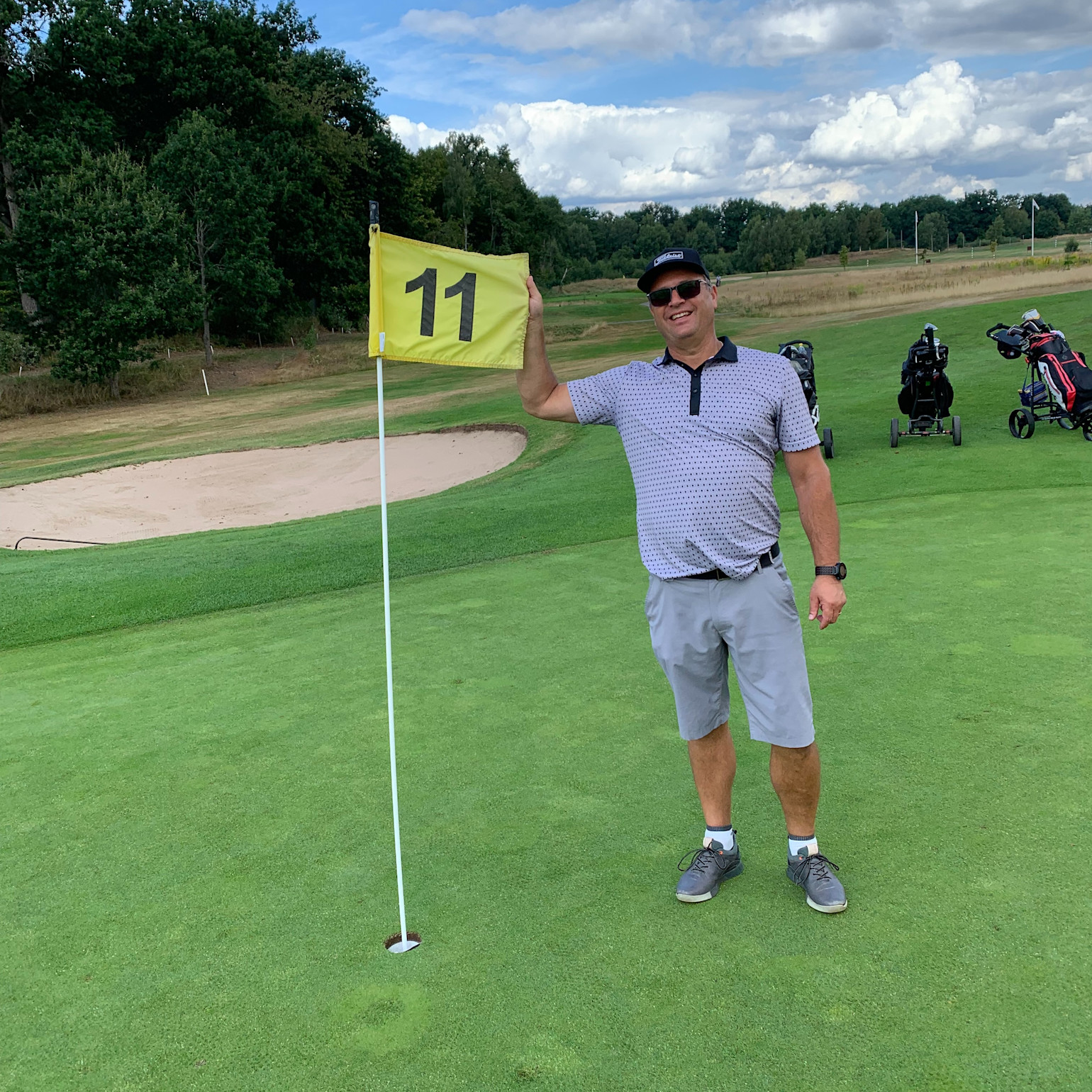 Mand i golfbeklædning står smilende ved hul 11 på en golfbane, holder flaget ved siden af greenen med golfbags i baggrunden.