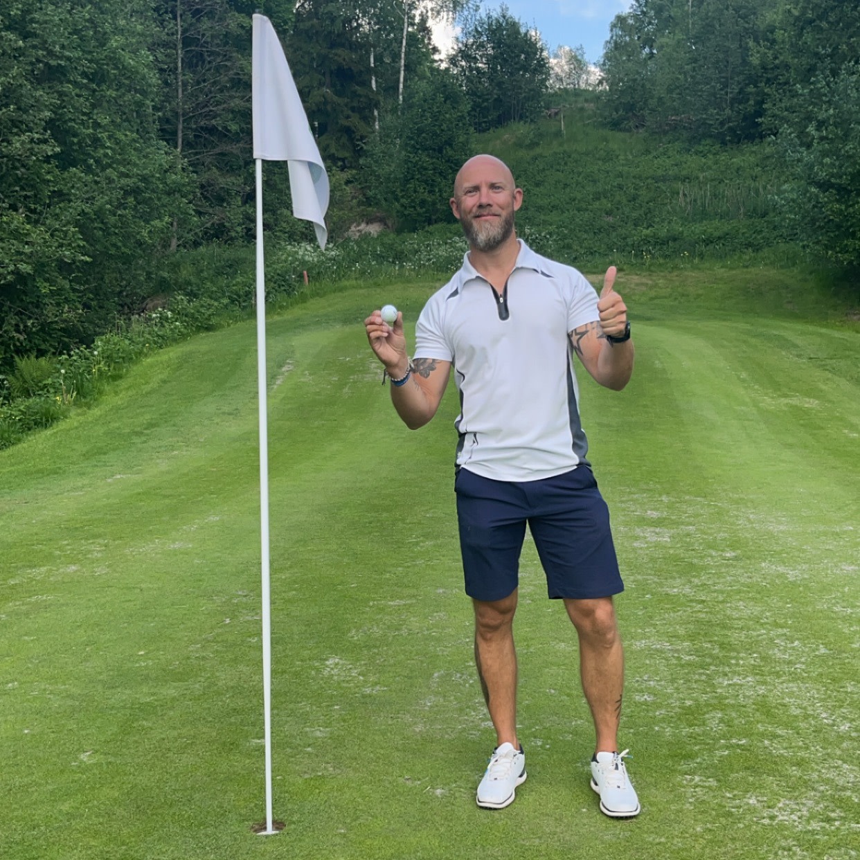 A man in golf attire holds a golf ball and gives a thumbs up next to a hole with a flag on a green golf course surrounded by trees.