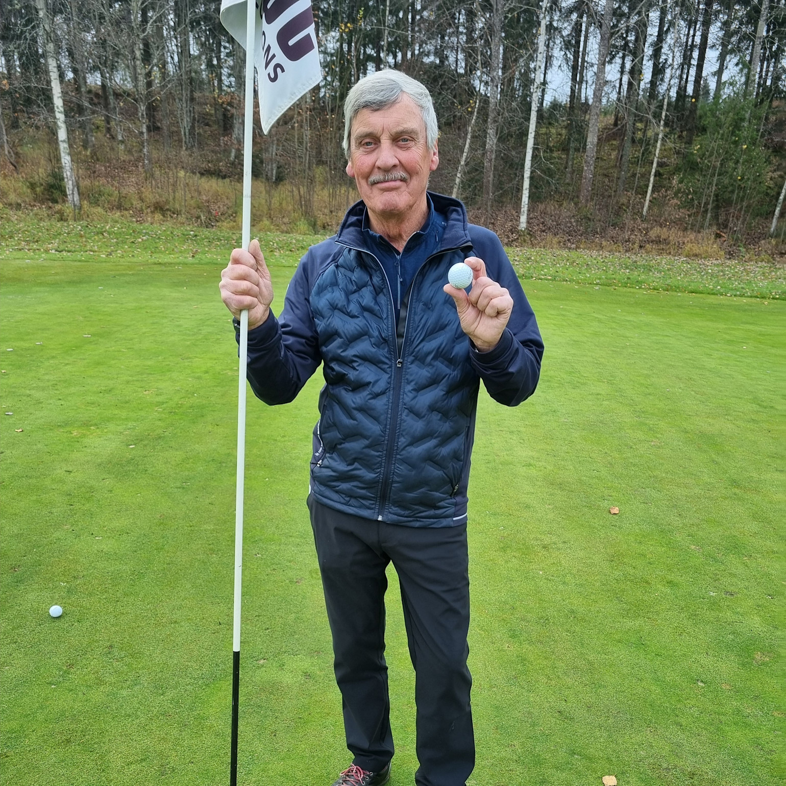Äldre man står på en golfbana, håller en flaggstång i ena handen och en golfboll i den andra, med träd i bakgrunden.