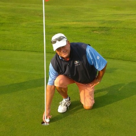 Mand iført kasket og solbriller bøjer sig ned ved et golfhul og samler en golfbold op på en grøn golfbane.