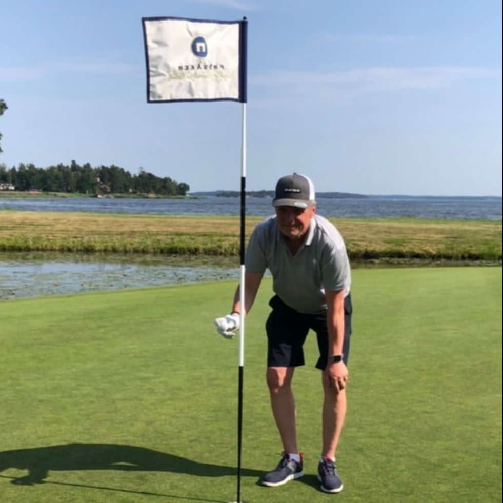 Mand iført grå polo og mørke shorts læner sig op ad en flagstang på en golfbane ved vandet på en solrig dag.