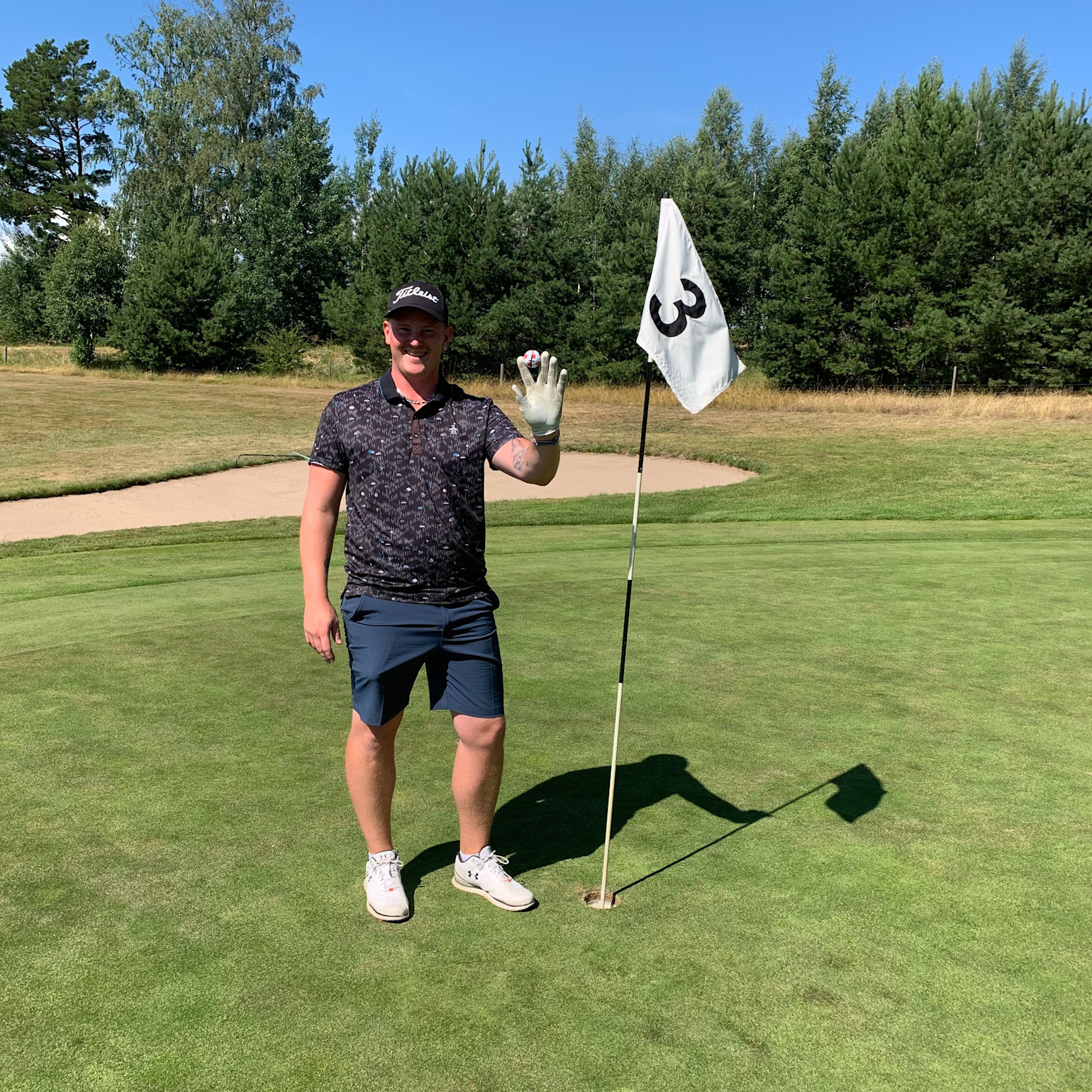 Mand iført golfbeklædning holder en golfbold op ved siden af flag nummer 3 på en golfgreen, med træer og blå himmel i baggrunden.