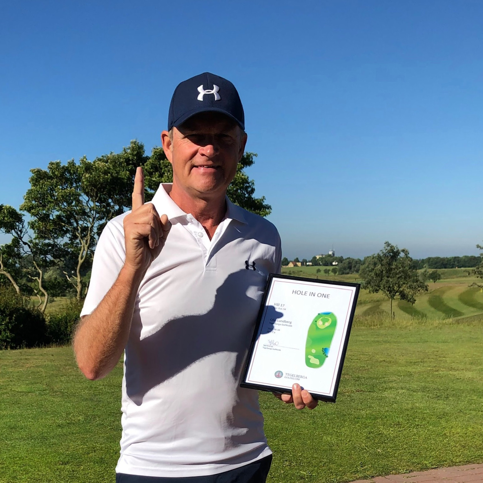 Mand i hvid polo og blå kasket holder et diplom med teksten "Hole in One" og viser én finger på en golfbane.