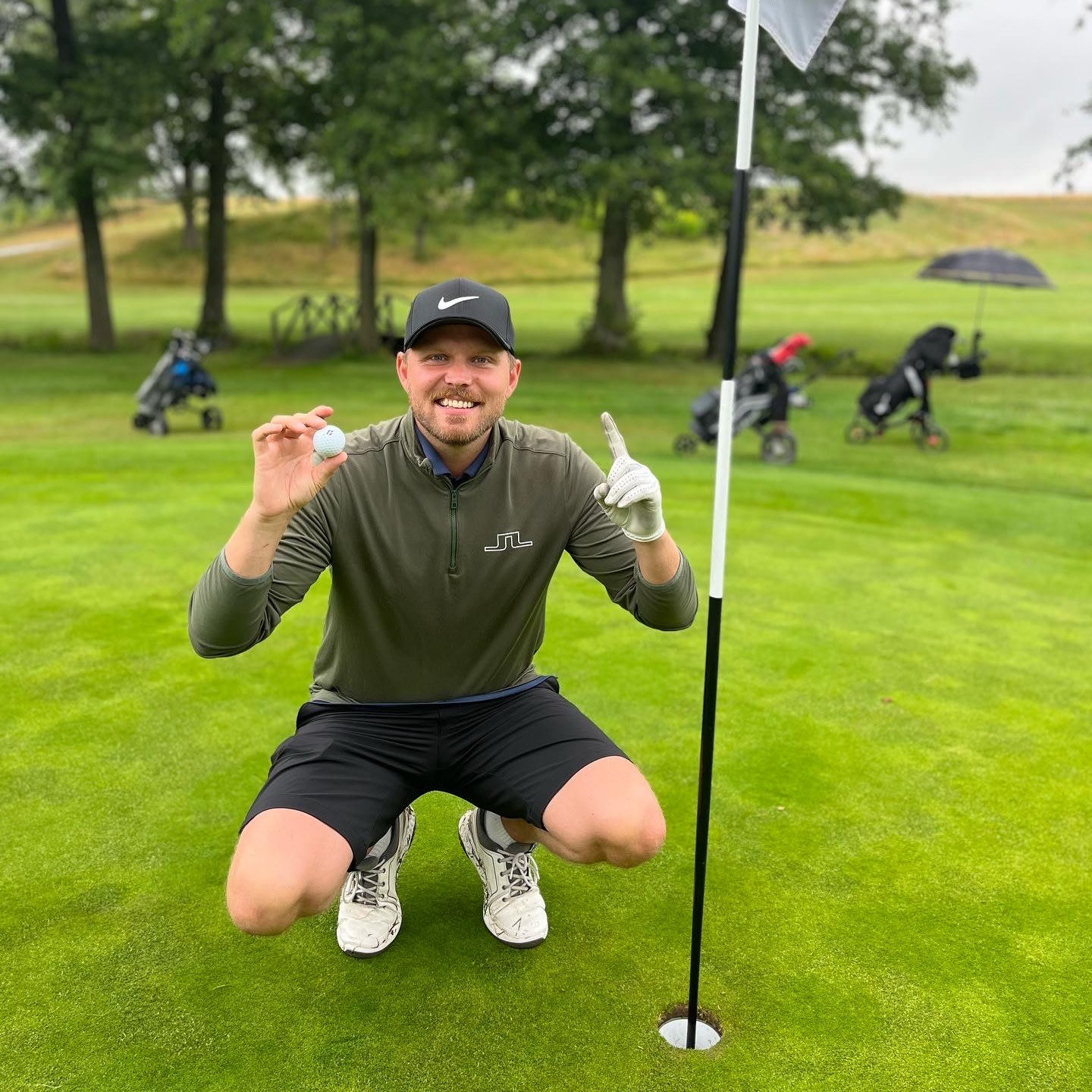 Mand i golfbeklædning knæler ved siden af et golfhul, holder en golfbold op og peger opad med en finger på en golfbane.