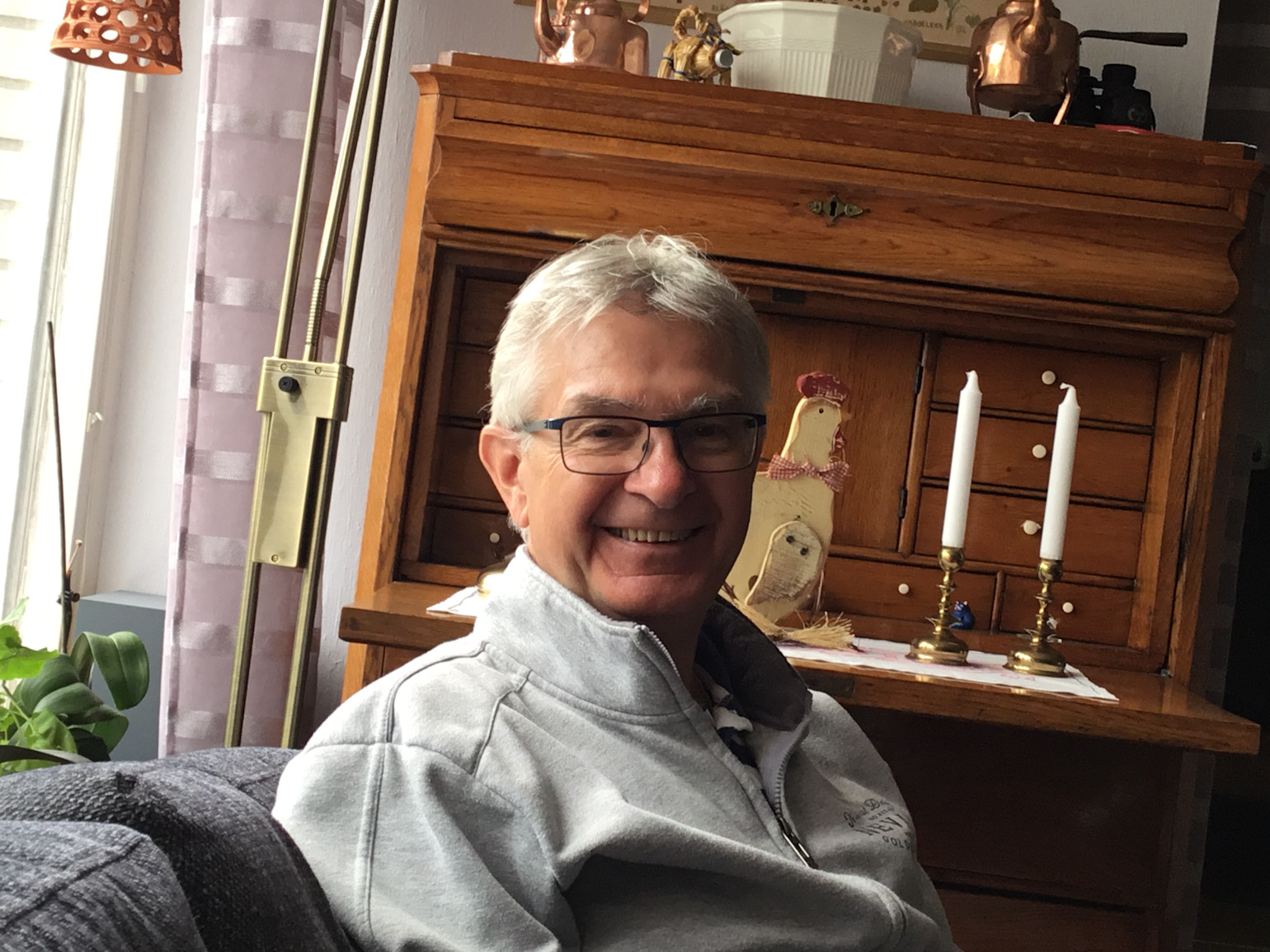 Smilende ældre mand med briller sidder i sofa foran et gammelt træskrivebord med to hvide lys og dekorationer.