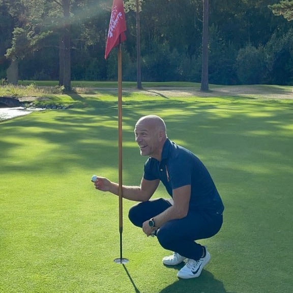 En mand iført golfbeklædning sidder på hug ved et golfhul og holder en golfbold ved siden af flagstangen på en solbeskinnet green.