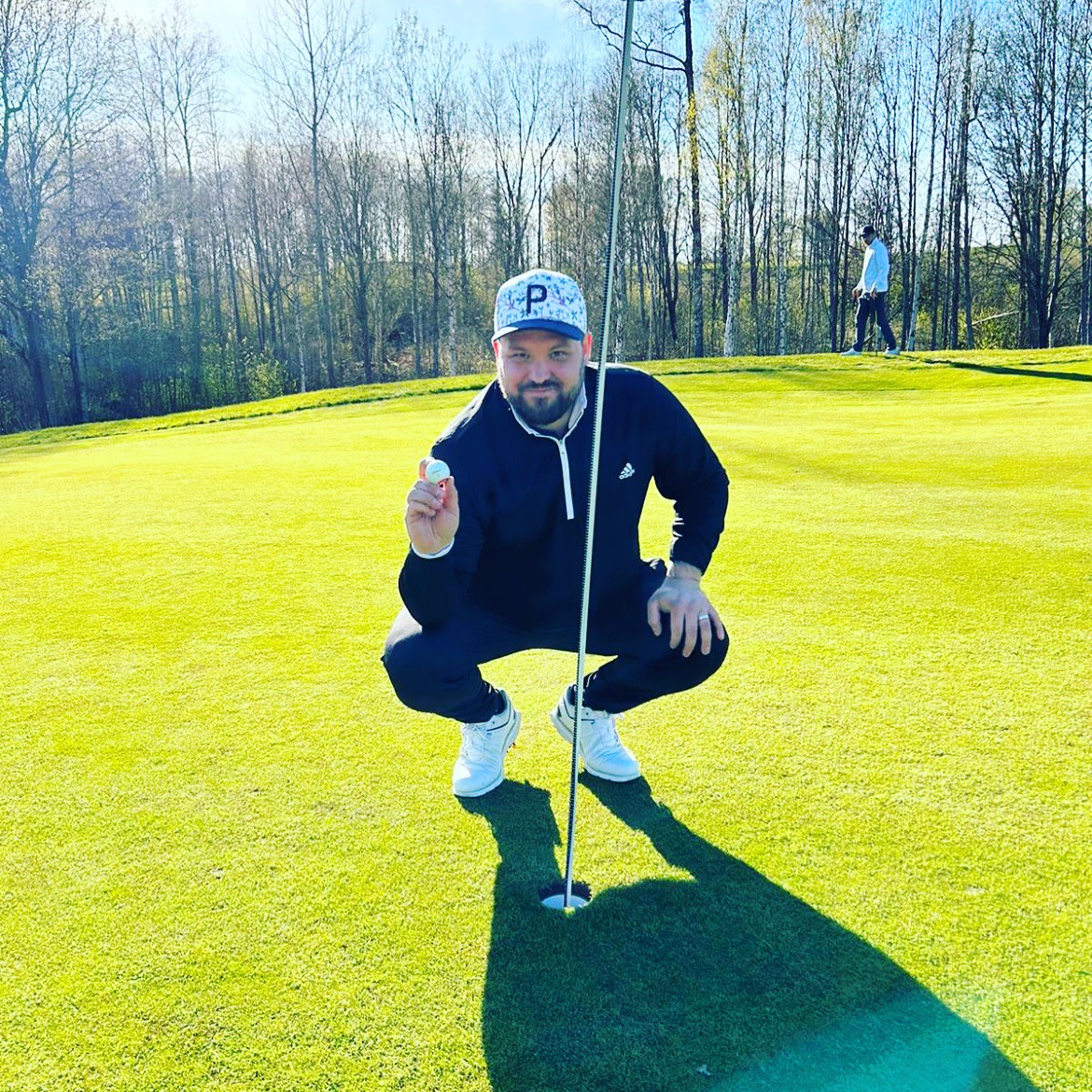 En mand i sporttøj sidder på hug ved et golfhul og holder en golfbold op ved siden af flagstangen på en solrig golfbane.