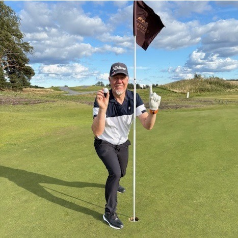 En mand i golfbeklædning smiler og poserer ved siden af et hulflag på en golfbane, mens han holder en golfbold op og peger opad med én finger.