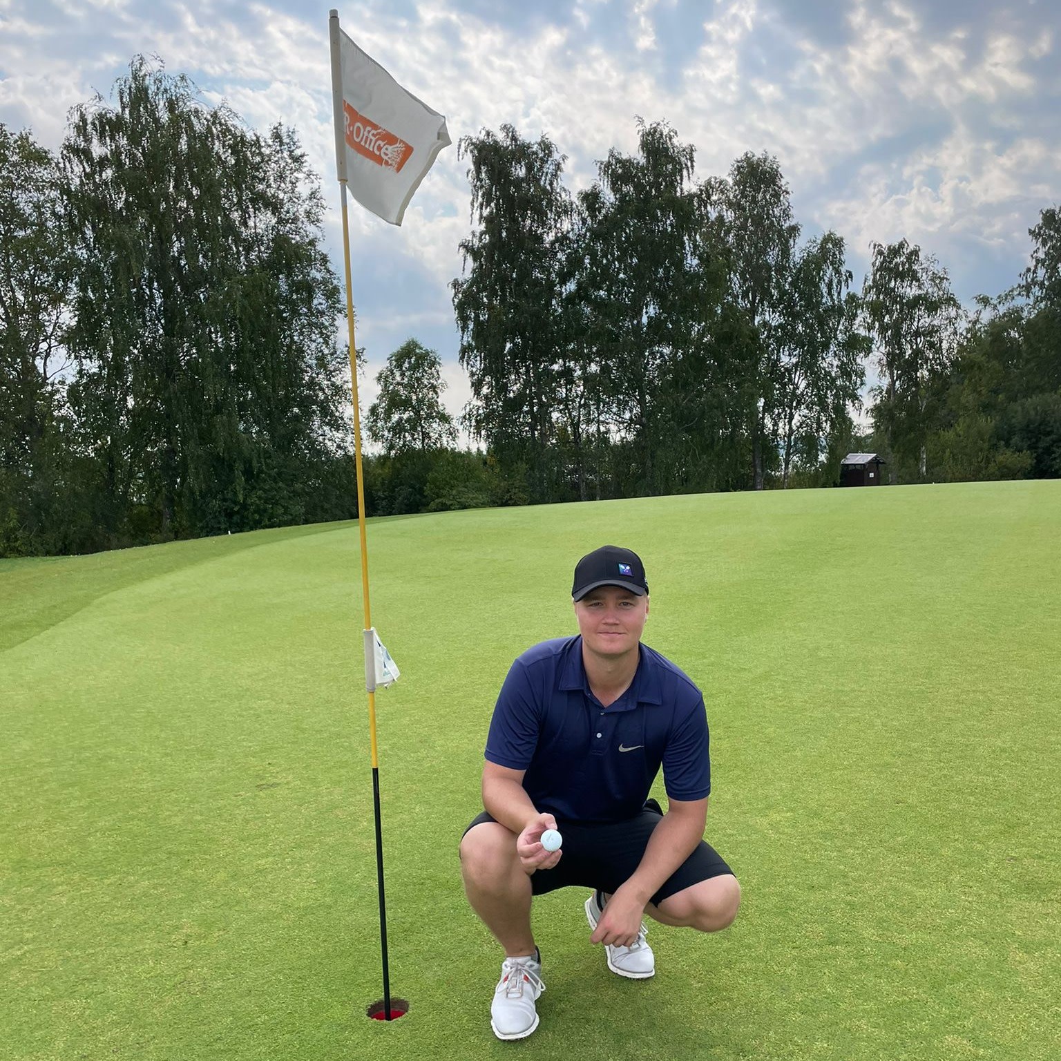 Mand iført blå polo og sort kasket sidder på hug ved et golfhul med flag, holder en golfbold på en grøn golfbane med træer i baggrunden.