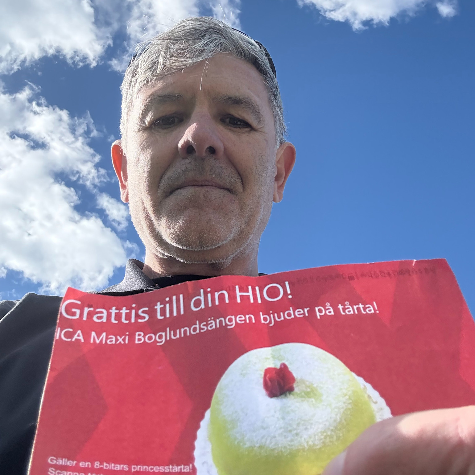 A person is holding a red card with the text "Congratulations on your hole-in-one! ICA Maxi Boglundsängen is treating you to cake!" against a blue sky.