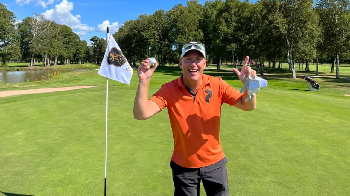Mand i orange polo-shirt holder en golfbold og smiler ved siden af et flag på en golfbane med træer i baggrunden.