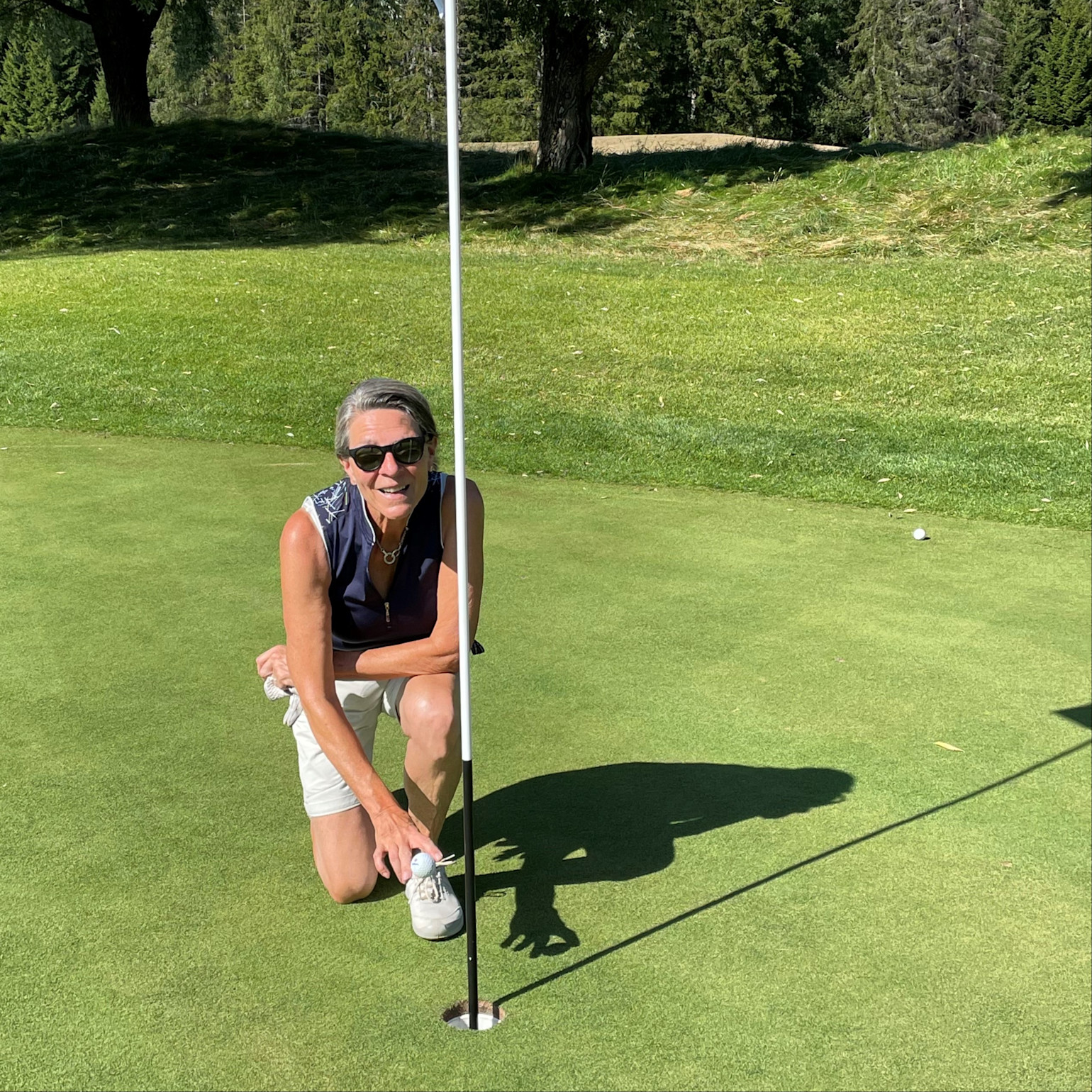 En person knæler ved et golfhul og holder en golfbold ved siden af flagstangen på en solrig green med træer i baggrunden.