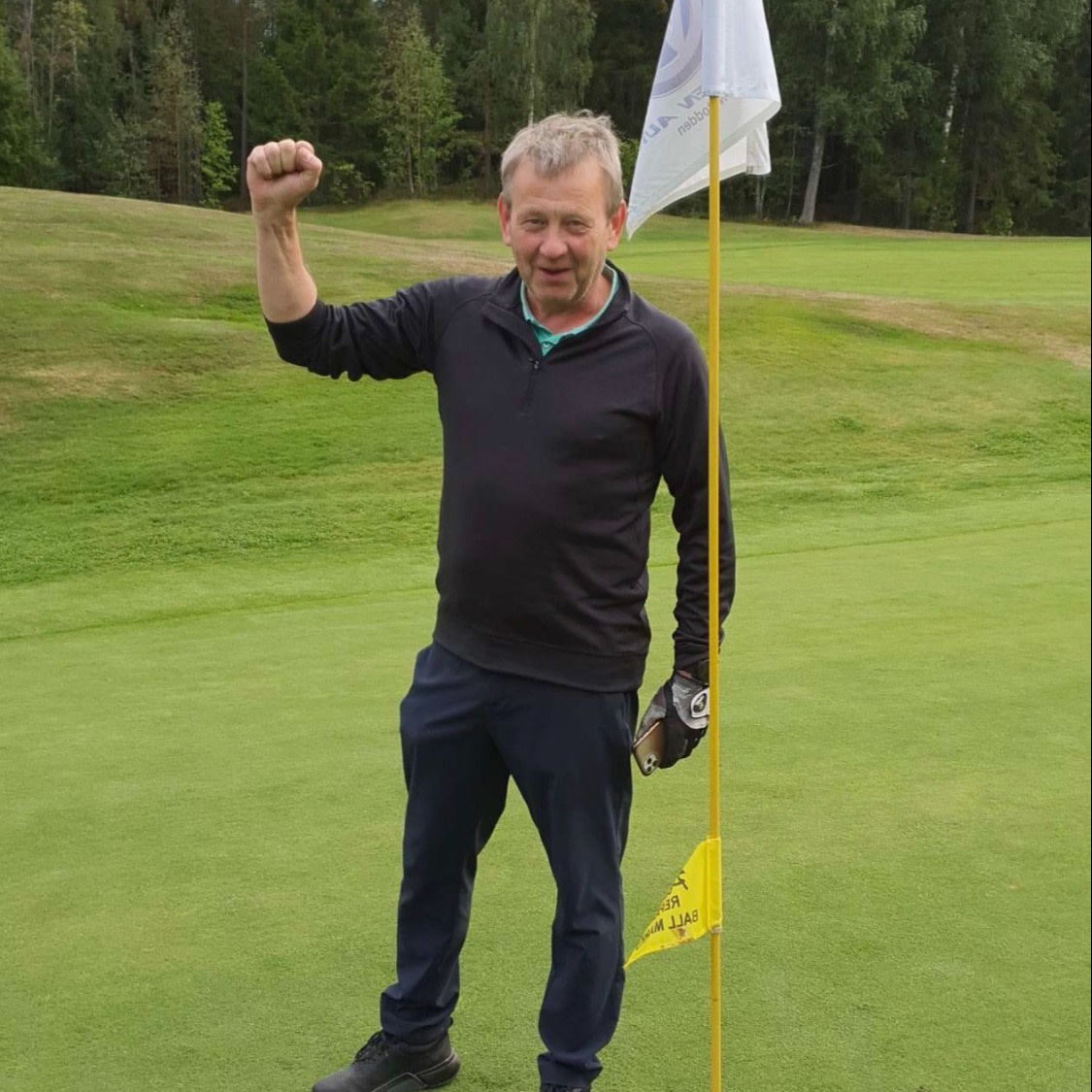 Man løfter sin knyttede næve i sejr ved siden af et golf-flag på en grøn golfbane med træer i baggrunden.