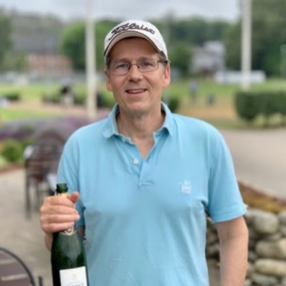 Mand iført lyseblå poloshirt og kasket holder en vinflaske udendørs i et parkområde.