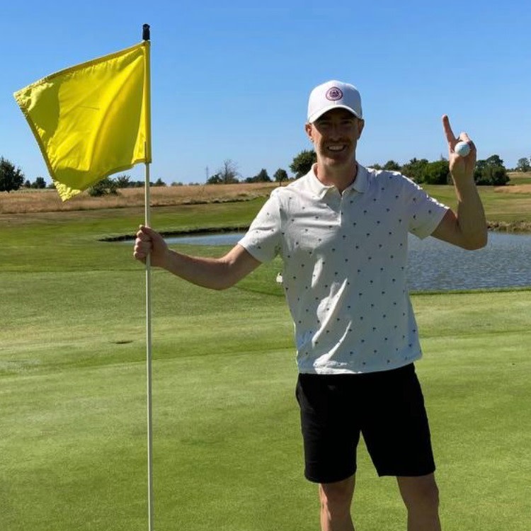 Man holder et gult flag og en golfbold på en golfbane, smiler og peger opad med en finger tæt ved en dam.
