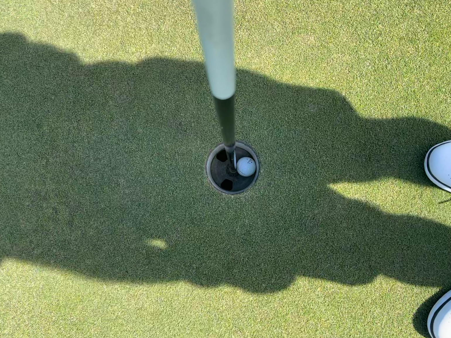 Golfbolden ligger i hullet ved siden af flagstangen på en puttinggreen, med to sko synlige og skygger på græsset.