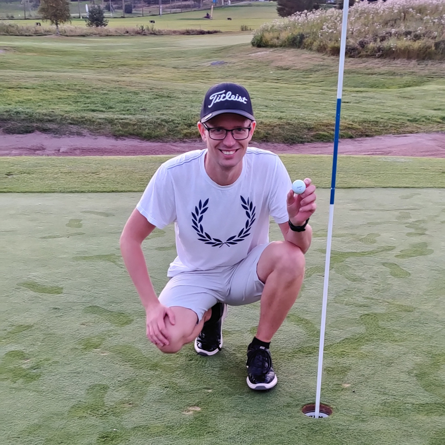 En mand i hvid t-shirt og kasket sidder på hug ved et golfhul på greenen og holder en golfbold op med et smil.