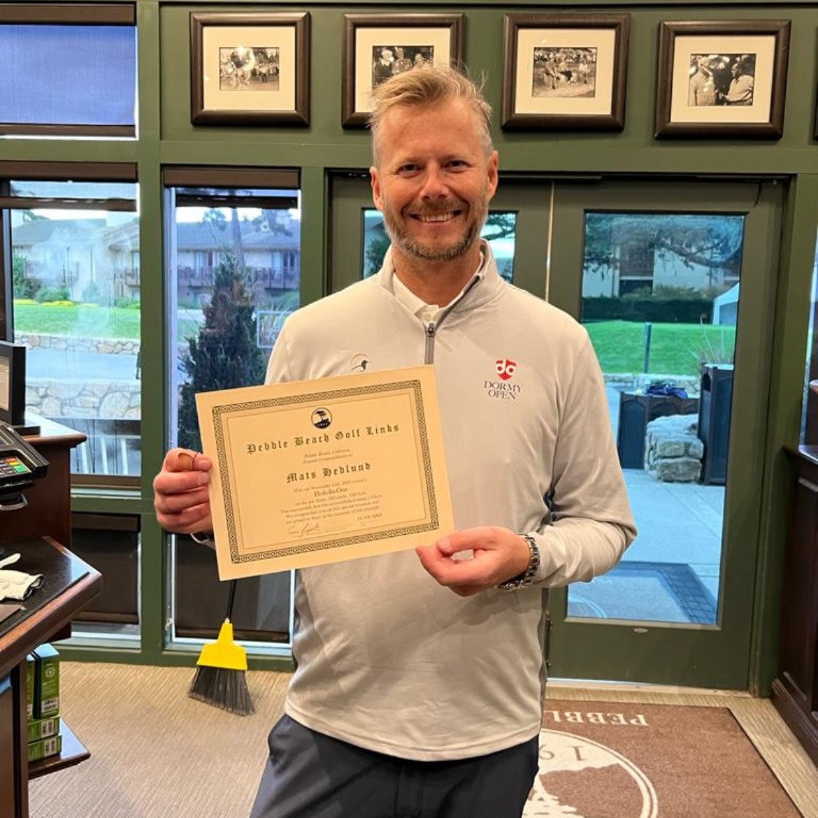 Man holder et diplom fra Pebble Beach Golf Links og smiler, står indendørs med vinduer og billeder i baggrunden.